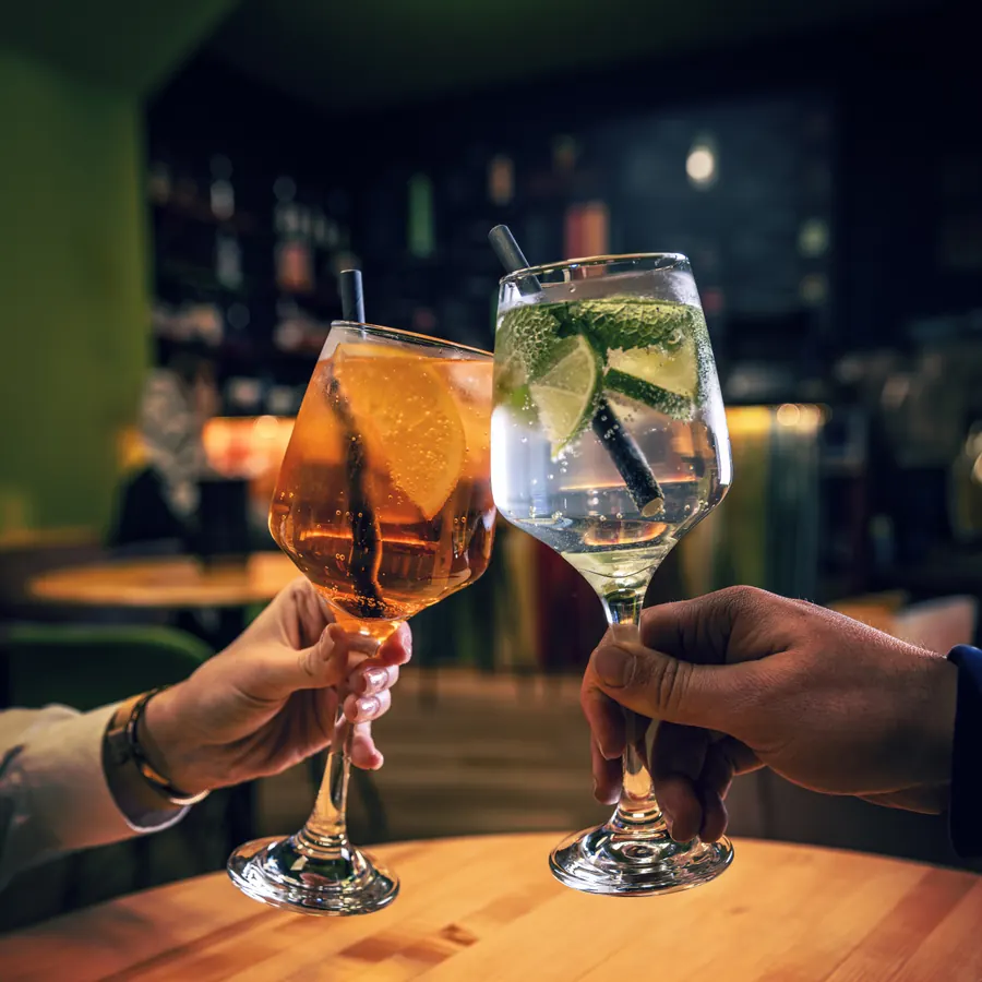 Dvoje gostiju nazdravlja spritzom i koktelom sa limetom za drvenim stolom